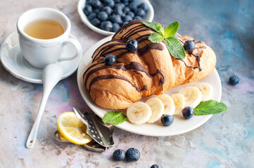 Croissant with chocolate, blueberries, bananas, and mint on a light grey background. Breakfast with a croissant and a cup of tea with lemon