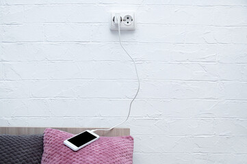 White smartphone lying on the pillow of bed connected to a charging electrical outlet.