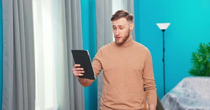 Bearded smiling tenage man using tablet for video conversation while relaxing in modern new gome. Concept of young people working at home.