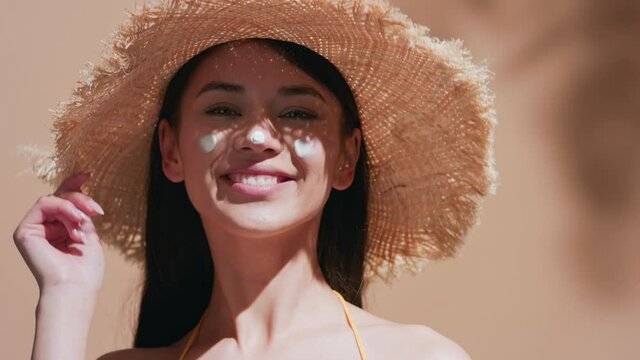 Young brown-haired asian woman in a straw hat applies some sunscreen on her nose, enjoys the sun and smiles for the camera against beige background |  Sunscreen commercial