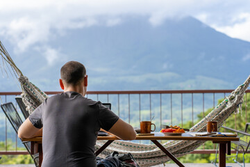 Man sitting on a table with breakfast while working with a view of a volcano in front of him