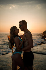 Fototapeta premium Beautiful couple in love on the background of the sunset by the sea. Young woman and man hugging by the sea at sunset