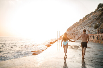 Fototapeta na wymiar A loving couple walks along the beach by the sea. Young family at sunset by the Mediterranean Sea. Vacation concept. A woman in a swimsuit and a man in shorts at sunset by the sea. Selective focus.