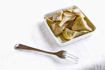 Artichokes hearts marinated with olive oil and herbs. Pickled artichoke with garlic in white plate on the table with white tablecloth. Homemade healthy eating.