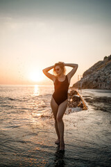 Fototapeta na wymiar Beautiful young woman in a black swimsuit alone on the beach by the sea at sunset. An adult slender girl resting on the ocean shore at sunset. Summer vacation concept. Selective focus