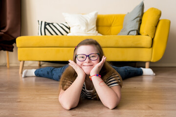 Portrait of smiling teenager girl with Down syndrome lying on the floor at home against the...