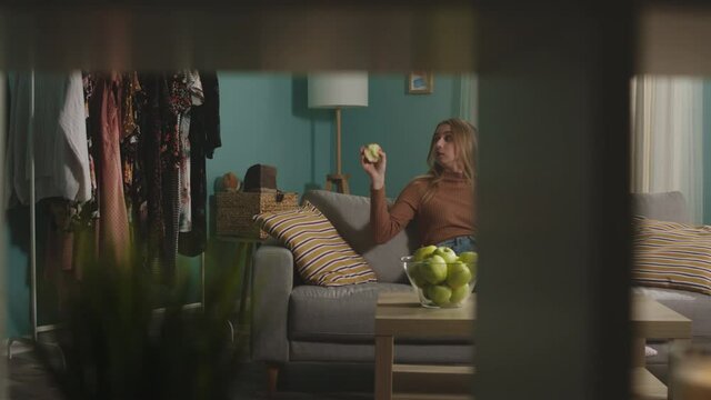 Girl in blue jeans and brown sweater sits on beige sofa on window background, bites red apple and talks on video call. On coffee table is glass jar with green apples. Camera dollies horizontally