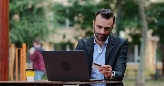 A man in a suit takes a bank credit card from the table, next to a laptop, and looks at it thoughtfully. Think before using bank cache.