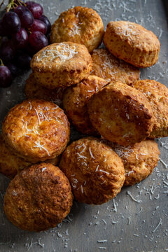 An angled view of a collection of cheese scones dusted with grated parmesan cheese. Placed on a grey background with red grapes to the top left of the image.