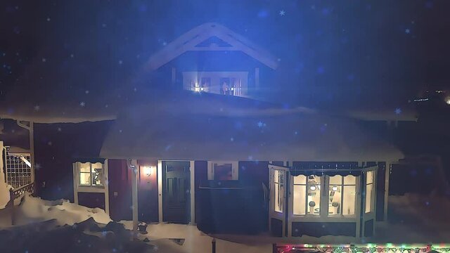 Video looks like 3D rendered cartoon of wooden house covered by much snow in countryside. Night, light snowfall, fly to window, away and up. Light rays, from lights and decorations. Cartoon effect
