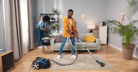 Fototapeta na wymiar African American young couple man and woman cleaning apartment, husband vacuuming carpet floor in living room while wife wiping dust disinfecting furniture in house, people moving in new flat