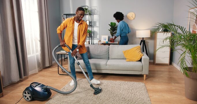 African American young couple man and woman cleaning apartment, husband vacuuming carpet floor in living room while wife wiping dust disinfecting furniture in house, people moving in new flat