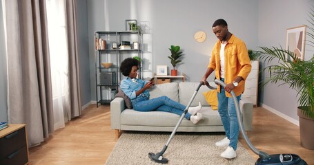 Happy handsome young African American husband mopping the floor in front while pretty wife resting on couch texting on smartphone browsing online social network app, cleaning home concept