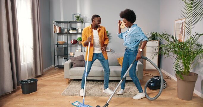 Joyful Happy African American Young Married Couple Wife And Husband Cleaning Living Room And Dancing, Guy Mopping And Woman Vacuuming Floor Having Fun Making Funny Rhythmic Moves In Cozy Modern Home