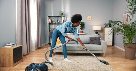 Young beautiful happy joyful African American woman cleaning modern living room vacuuming carpet and dancing having fun playing vacuum cleaner like guitar, housekeeping concept, clean up - obrazy, fototapety, plakaty