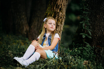 Portrait of a beautiful little girl with ponytails sitting on the ground, leaning against a tree in a summer park by the lake