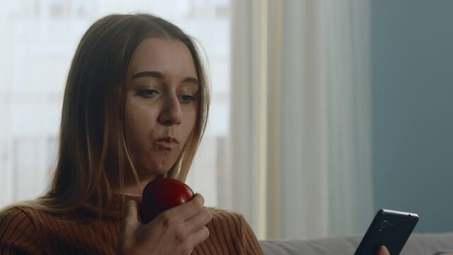 Young woman in brown sweater sits on beige sofa on window background, biting red apple and looking at something on phone. Close-up.
