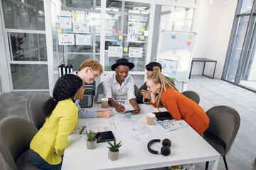 Obraz premium Group of multiracial smiling confident office coworkers, sitting at the table and having a break after meeting, laughing while looking on cell phone screen