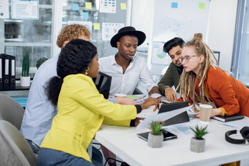 Team of likable creative high-skilled diverse young colleagues, working together over joint project and analyzing different diagramms and infographics during meeting in modern office