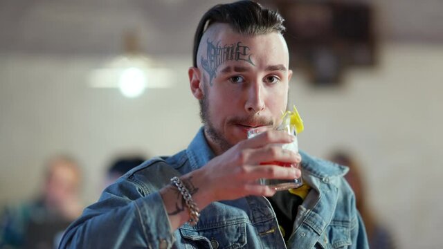 Handsome confident young man with tattoo on face and piercing drinking cocktail in slow motion, clinking with camera and smiling. Portrait of happy relaxed Caucasian millennial posing in bar.