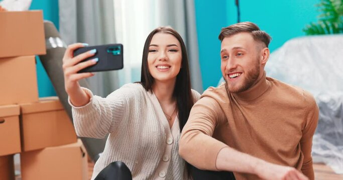 Beautiful caucasian young couple taking selfie and shooting video on cell phone sitting on wooden floor in new apartment.