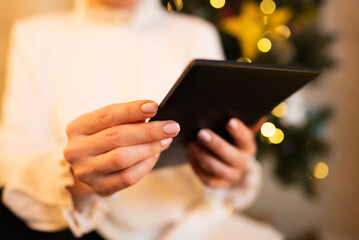 Woman in white reading interesting e-book reader while sitting near Christmas tree at home. Cozy winter pic of lady relaxing alone with novel.
