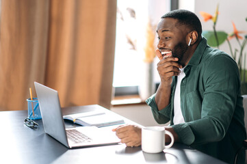Joyful African American guy dressed in stylish clothes uses a laptop for online work, sitting at his desk, happily looks at the screen, got a good message