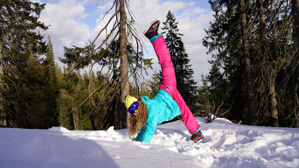 Bright girl in a ski suit performs yoga on snow in forest. flexible athlete warm-up before...