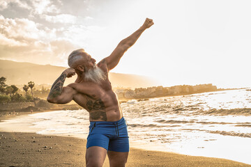Fototapeta na wymiar Age is just a number. In a healthy body, healthy mind. Senior man with white stylish beard showing his muscular fit body with tattoos on the beach.