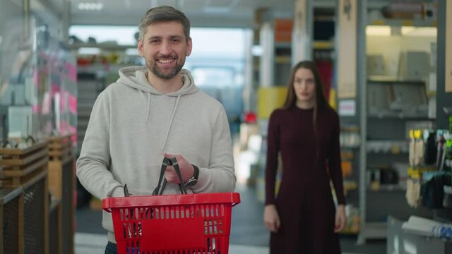 Portrait of satisfied Caucasian man looking at camera and smiling as irritated woman yelling and gesturing at background. Happy male shopper posing in hardware store with annoyed wife shouting.