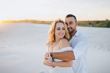 A young bearded man hugs a blonde against a background of white sand and smiles. Portrait of emotional people at sunset. Love of newlyweds in the desert. Love story.