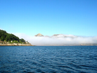 A bank of morning fog floating over water, Islac Cies (Cies Islands) Galicia, Spain, Europe; long view from an island towards the mainland, peaks of mountains visible above the cloud.