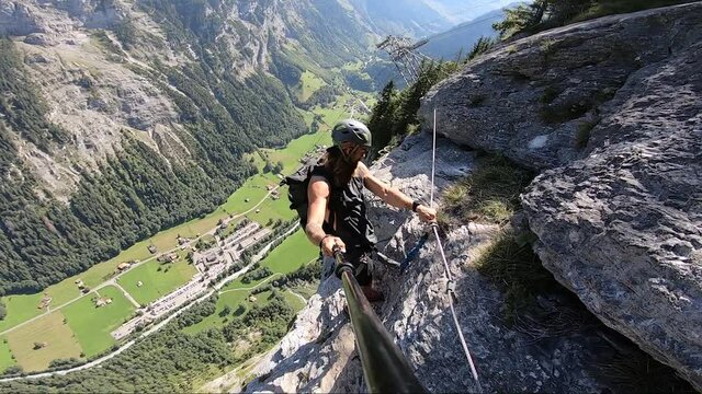 Young man takes a selfie video as he stands on the very narrow and dangerous path on Via Ferrata route in Switzerland with the gorgeous and green valley below during a hot clear summer day.