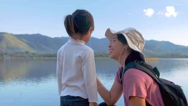 Happy mother and little daughter listen to music with smartphone earphone together on beautiful sunset lake background. Family lifestyle and Vacation concepts.