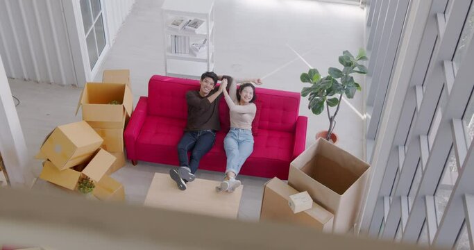 High angle, slow motion - Happy young Asian couple tenants give a high five on the sofa and look at camera after unboxing on house moving day.