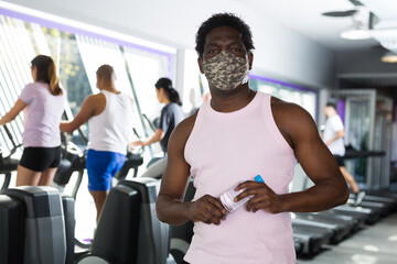 Positive man wearing face mask for viral protection resting after exercises in fitness gym holding drinking water