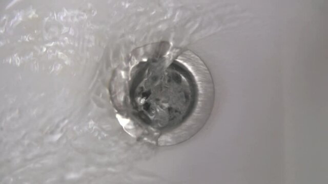 Water flows from the tap into the bathtub and streaming down the metal drain hole. Running water and bathroom sink drain slow motion close up.