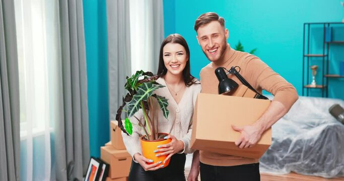 Portrait of smiling couple standing at new home carrying boxes. Portrait of happy married couple in new apartment looking at camera smiling holding in hands box and flowerpot.