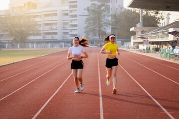 Two young Asian women in sports outfits jogging on running track in city stadium in the sunny morning to keep fitness and healthy lifestyle. Young fitness women run on the stadium track