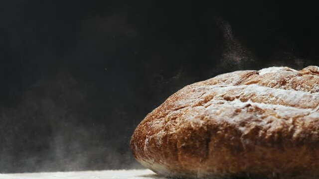 Loaf of healthy fresh rye bread covered with white flour falls down on table on black background extreme close view slow motion