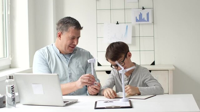 Father and son sitting at table in home office playing and working with two models of wind turbine