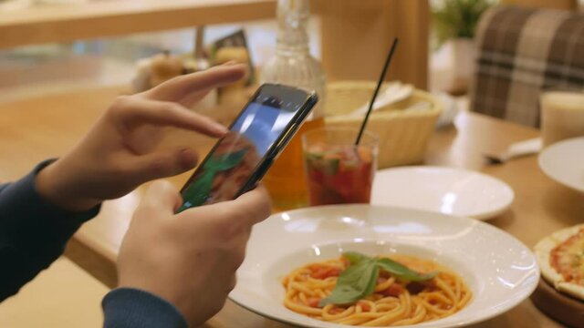 A visitor is sitting at a table in the restaurant. Various dishes are on the table. Italian pasta in sauce with basel leaves, berry cocktail and pizza. A man photographs food on a smartphone.Close-up.