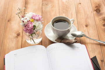 A view of a workplace with pink and white flowers in a small glass jar of water, as well as a red notebook with a pen and coffee on a wooden table.