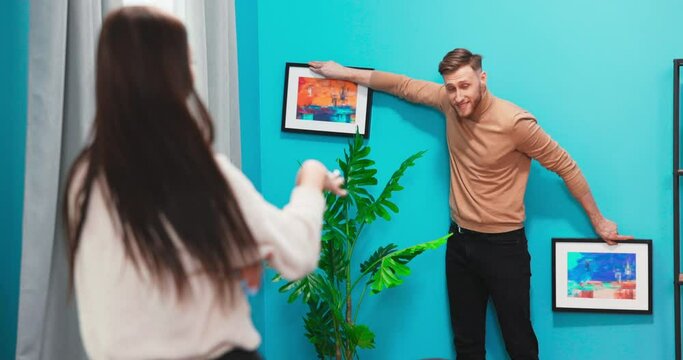 Happy Couple Hanging Picture on the Wall, Boyfriend Moves It, Girlfriend Tells Him when the Frame is Hanging Straight. Funny Moment in Young Couple's Life. Modern Stylish Apartment.