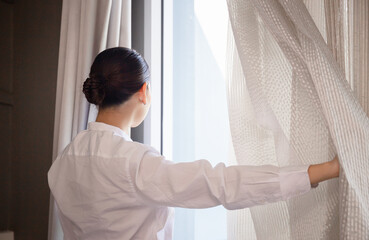 Young woman standing by bedroom window and opening curtains, girl open window curtains
