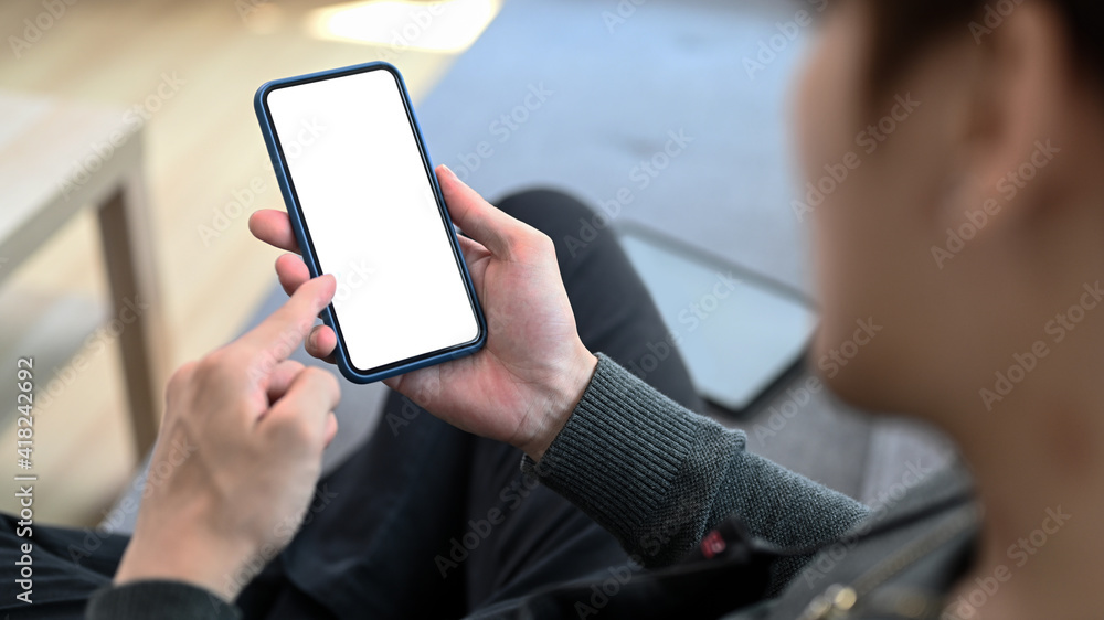 Poster close up view of young man holding mobile phone with empty screen while sitting on sofa at home.
