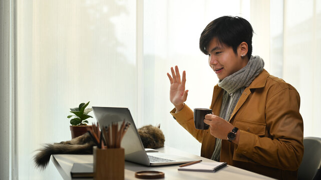 Asian man having video call on laptop computer and greeting his colleagues.