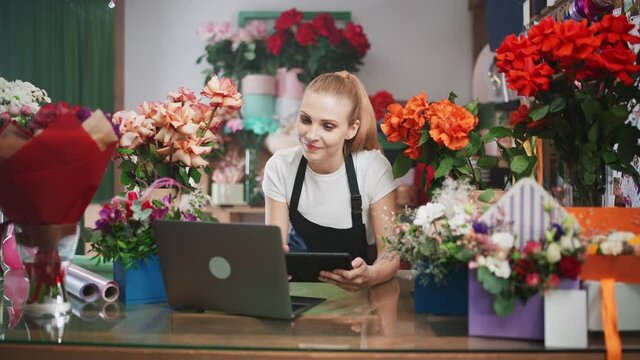 Florist woman discusses the order by talking on video call, using a laptop and screen tablet in a flower shop, working online.