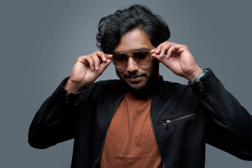 Obraz premium Stylish hindu hipster wearing fashionable clothing poses in gray background taking off his sunglasses.
