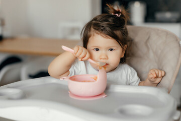 The child himself eats food at home in the kitchen. Cute little Asian girl eats fruit puree. The child is sitting in a high chair. The girl eats with a spoon. The child himself eats food